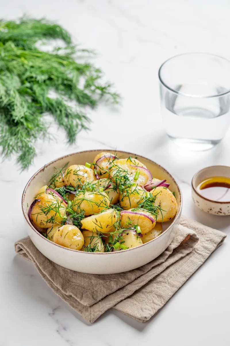 Erdäpfelsalat in einer Schüssel, daneben frische Kräuter und ein Kännchen Öl