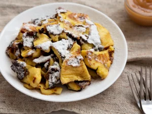 Kaiserschmarrn mit Staubzucker auf einem Teller, dahinter ein Glas Apfelmus