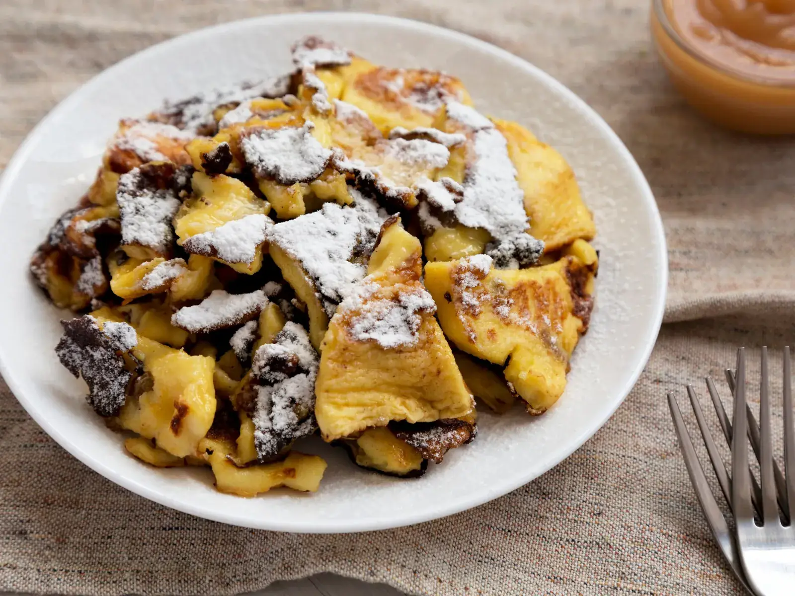 Kaiserschmarrn mit Staubzucker auf einem Teller, dahinter ein Glas Apfelmus
